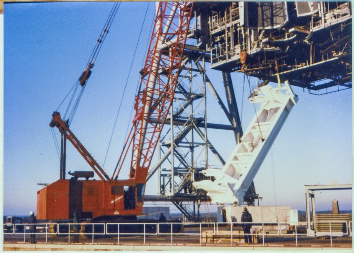 The PHGM Bridge Beam has been rotated and tilted to its final lifting orientation, and the main sense of the lift is now underway as it rises toward the support rails in the uppermost reaches of the space inside the Payload Changeout Room which are already in place, waiting to bear its load.